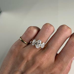 Pu&ograve; includere: Un anello d'oro con tre diamanti taglio ovale disposti in fila, affiancato da una fascia d'oro pi&ugrave; piccola. I diamanti brillano. L'anello &egrave; mostrato su un dito.
