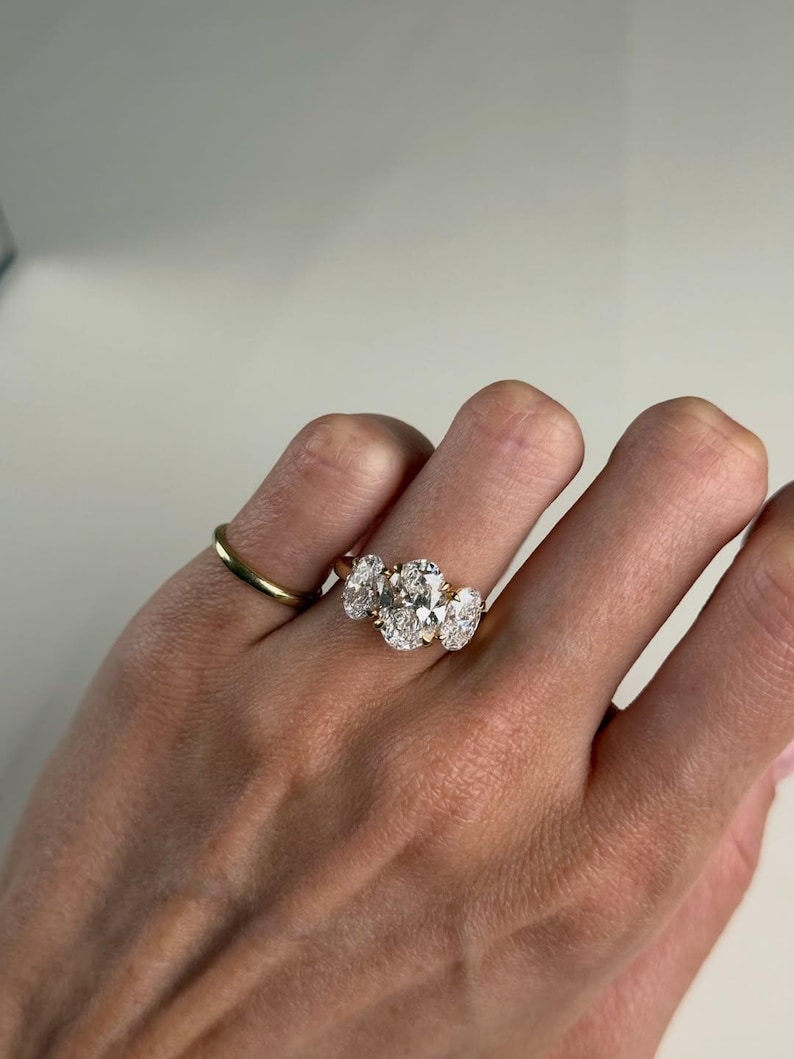 Pu&ograve; includere: Un anello d'oro con tre diamanti taglio ovale disposti in fila, affiancato da una fascia d'oro pi&ugrave; piccola. I diamanti brillano. L'anello &egrave; mostrato su un dito.
