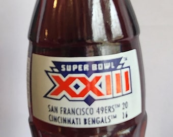 Puede incluir: Una botella de vidrio vintage llena de un líquido oscuro, probablemente un refresco. La botella presenta una etiqueta del Super Bowl XXIII, con los logotipos de los 49ers de San Francisco y los Bengals de Cincinnati. La tapa es metálica.
