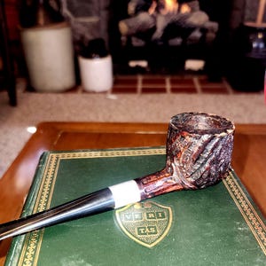 Pode incluir: Um cachimbo de tabaco esculpido com uma tigela castanha escura e um bocal preto, pousado em um livro verde. O cachimbo tem uma faixa prateada e está situado em um fundo aconchegante com uma lareira e um copo de líquido vermelho.
