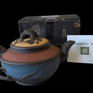 May include: A multi-colored clay teapot with a blue base, a reddish-brown midsection, and a tan lid featuring a sculpted branch handle. The teapot is accompanied by a black box and a white card labeled KingWare teapots with a logo.