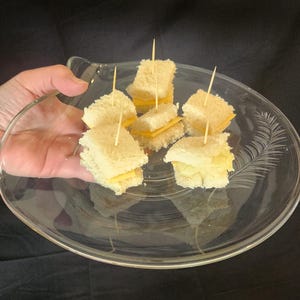 May include: A clear glass serving plate with a decorative leaf pattern holds several small sandwiches. Each sandwich is made with white bread and cheese, secured with a toothpick. The plate is held in a person's hand against a dark background.