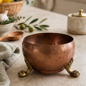 May include: A round copper bowl with a smooth, weathered finish, supported by three ornate brass feet shaped like animal claws. The bowl has a deep, warm metallic tone with visible dark spots and a wide, open top design for holding items.