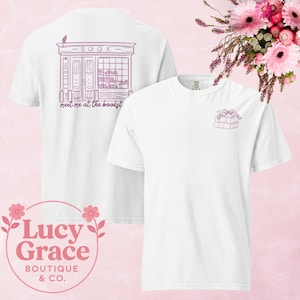 Könnte beinhalten: Weißes T-Shirt mit einer Buchladen-Grafik auf dem Rücken und einem Stapel Bücher auf der Vorderseite. Das Buchladen-Design enthält das Wort "BOOK" und den Satz "meet me at the bookstore". Auf der Vorderseite des T-Shirts befindet sich ein kleiner Stapel Bücher mit einer Schleife.