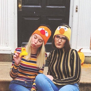 May include: Two people wearing handmade crochet hats with cartoon character designs. One hat is orange with a red beak and white eyes, the other is yellow with an orange beak and white eyes. One person holds a yellow rubber duck.