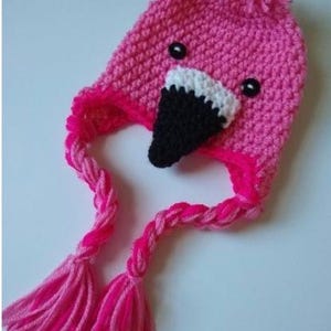 May include: A bright pink crocheted hat shaped like a flamingo, featuring a black and white beak, two small black button eyes, a yarn pom-pom on top, and two long braided earflaps ending in thick pink yarn tassels.