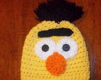 May include: A yellow crocheted hat with a black tuft of hair on top. The hat features large white eyes with black button pupils and an orange beak. The hat is designed to resemble a cartoon character.