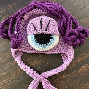 May include: A handmade crochet hat in shades of purple, featuring a large eye design. The hat has a braided chin strap with tassels and a textured, curly hair design around the top. The eye is detailed with black, white, and blue accents.