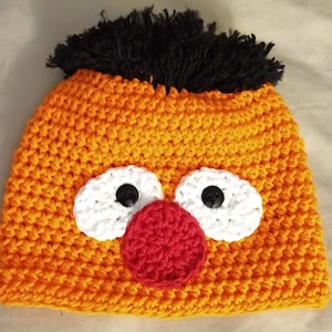 May include: Handmade orange crochet hat with white eyes, black button pupils, a red nose, and black fringe hair. This whimsical hat is perfect for costume parties or everyday wear.