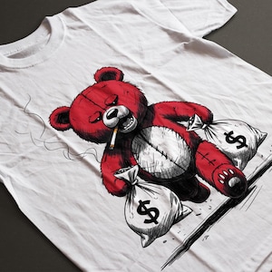 May include: White t-shirt featuring a red teddy bear smoking a cigarette, with a stitched belly and holding two money bags with dollar signs. The graphic is a detailed illustration.