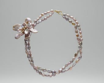 May include: A double-strand necklace featuring oval-shaped pearls in shades of lavender and gray. A flower-shaped pendant with gold accents and pearl details adorns the necklace. The necklace is suspended against a neutral background.