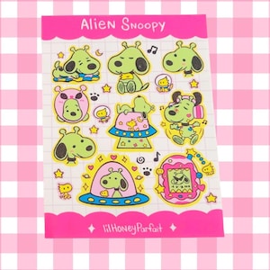 May include: A sheet of stickers featuring cartoon Snoopy characters with alien-themed designs. The stickers are primarily green, pink, and yellow, with the text "Alien Snoopy" at the top. The sheet has a pink scalloped border and the text "lilHoneyParfait" at the bottom.
