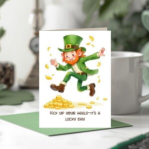 Puede incluir: Tarjeta de felicitación con un duende de dibujos animados vestido de verde, saltando sobre monedas de oro. La tarjeta dice "Kick up your heels—it's a LUCKY DAY". El fondo es blanco, con un sobre verde.