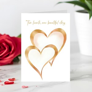 May include: A white greeting card with two overlapping gold-colored hearts. The card features the text "Two hearts, one beautiful story" in gold script. A red rose and heart-shaped candies are visible in the background.