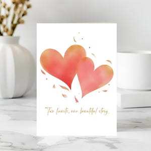 May include: A greeting card featuring two watercolor hearts in shades of pink and orange. The card has the text "Two hearts, one beautiful story" in gold script. The card is on a white background.
