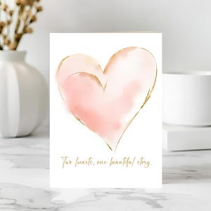 May include: A greeting card with a watercolor-style pink heart design outlined in gold. The card features the text "Two hearts, one beautiful story" in gold script. The card is on a white background.