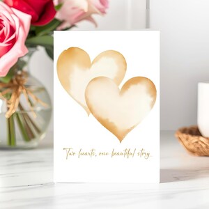 May include: A white card features two watercolor-style hearts in shades of gold. The phrase "Two hearts, one beautiful story" is written in gold script below the hearts. The card is displayed near a vase of roses.