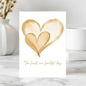 May include: A greeting card with a watercolor design of two overlapping hearts in shades of gold and tan. The card has the text "Two hearts, one beautiful story" in gold script.