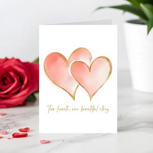 May include: A white greeting card featuring two overlapping pink hearts with gold outlines. The card has the text "Two hearts, one beautiful story" in gold script. A red rose and decorative hearts are in the background.