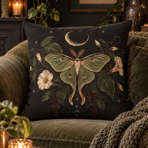 May include: A square throw pillow featuring a black background with a large, central green moth illustration. The design includes white morning glory flowers, dark green leaves, and a golden crescent moon symbol positioned above the moth.