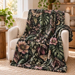 May include: A soft throw blanket featuring a dark background with a botanical print of large green monstera leaves, pink flowers, and butterflies. The design includes small golden crescent moons and stars scattered throughout the pattern.