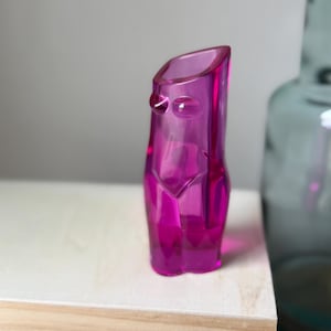 May include: A vibrant magenta-colored glass vase with a unique, abstract form. The vase has a sculpted shape with rounded details and a slanted opening. The design is reminiscent of a human form. The vase is displayed on a light-colored wooden surface.