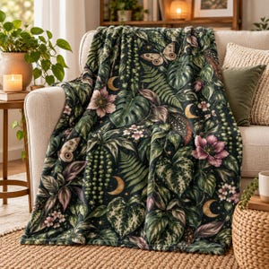 May include: A dark green throw blanket featuring a repeating pattern of monstera leaves, ferns, string of pearls, pink flowers, butterflies, and golden crescent moons. The soft fabric drapes over a light-colored sofa, displaying a detailed botanical and celestial print.