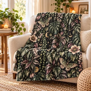 May include: A plush throw blanket featuring a dark background with a botanical pattern of green monstera leaves, trailing vines, light-colored flowers, and butterflies. The design includes small golden crescent moon and star motifs.