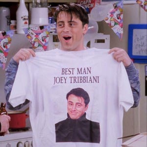 May include: White t-shirt with the text "BEST MAN JOEY TRIBBANI" above a smiling image of a man. The man is holding the shirt up, with a surprised expression. The shirt is a crew neck style.