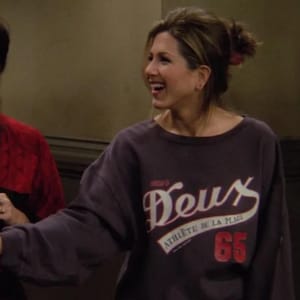 以下が含まれることがあります： 白い筆記体で「Deux」という文字、白で「Athlète de la Plage」という文字、赤で「65」という数字が入った濃いグレーのスウェットシャツ。 スウェットシャツは人が着ています。