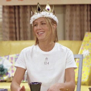 May include: A woman wearing a gold and white crown and a white t-shirt with the number 12-4 printed on it. The crown has gold and black accents. The woman is smiling and sitting at a table.