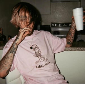 May include: A light pink t-shirt featuring a cartoon character design with the text "HELL BOY" in black. The shirt is worn by a person with tattoos and a white cup in their hand.
