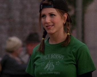 May include: A green t-shirt with the text "GIRLS BOXING BRONX NEW YORK" and a black baseball cap. The shirt has a graphic of boxing gloves and stars. The person is smiling.