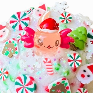 May include: A round, light pink object covered in white, green, and red sprinkles and decorative elements. The decorations include miniature gingerbread houses, Santa faces, snowflakes, candy canes, and red and green peppermint candies. A cartoon character wearing a Santa hat is in the center.