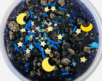 May include: A clear container filled with a dark blue slime with black rocks, yellow crescent moons, blue and yellow stars, and blue glitter.