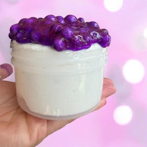 Pode incluir: Um frasco de plástico transparente cheio de slime branco, coberto com uma substância roxa vibrante e texturizada. O frasco é segurado em uma mão contra um fundo rosa suave com bokeh branco.