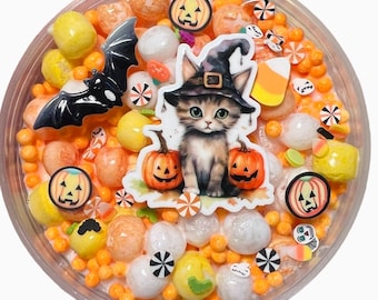 May include: A clear container filled with orange and yellow candy, decorative shapes, and a sticker of a cat wearing a witch hat and holding pumpkins. A black bat and pumpkin stickers are also included. The candy is a mix of sizes and shapes.