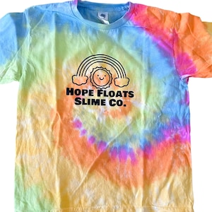 Pode incluir: Uma camiseta tie-dye com um arco-íris e um sol sorridente. O texto "HOPE FLOATS SLIME CO." está impresso abaixo do gráfico.