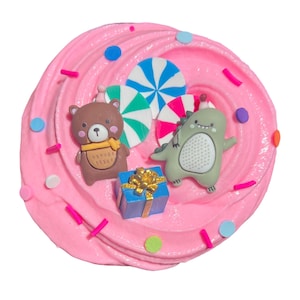 Pode incluir: Slime rosa com elementos decorativos. Uma figura de urso castanho com cachecol amarelo, uma figura de dinossauro verde, uma caixa de presente azul com laço dourado e doces coloridos estão dispostos no slime. O fundo é branco.