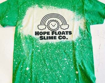 May include: Green t-shirt with a bleach-splattered design. The shirt features a black and white graphic of a rainbow, sun, and the text "HOPE FLOATS SLIME CO."