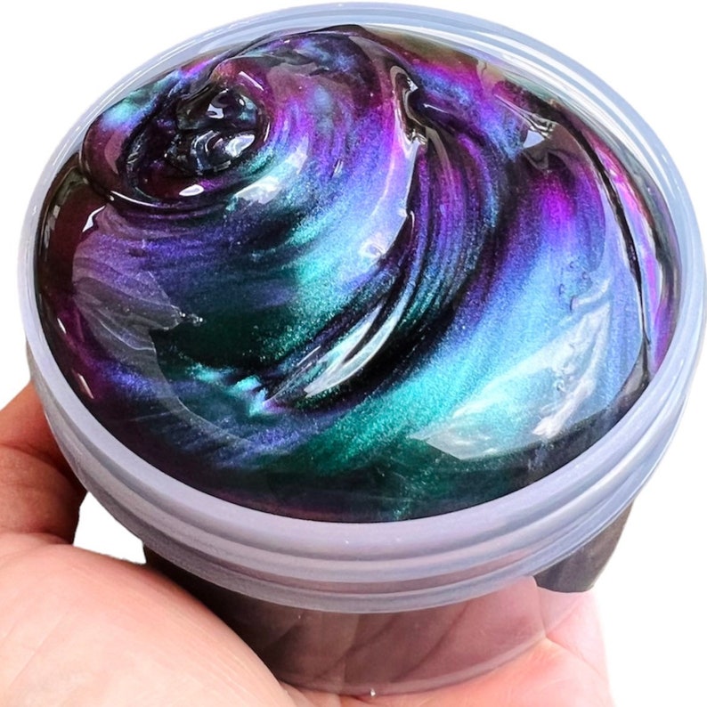 May include: A clear plastic container filled with iridescent slime. The slime is a dark green, purple, and black color with a swirling pattern.
