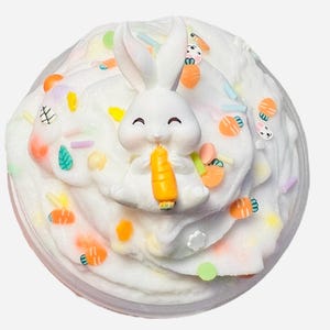 May include: A white, fluffy slime with a smiling white bunny figurine holding an orange carrot. The slime is decorated with colorful sprinkles, carrot and bunny-shaped embellishments, and other small, colorful shapes. The slime is in a clear plastic container.