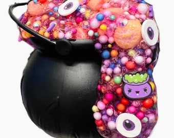 May include: A black plastic cauldron filled with purple and pink textured slime. The slime is overflowing with colorful foam beads, round sprinkles, and flat circular eye charms. A small purple cauldron-shaped charm is embedded in the slime.