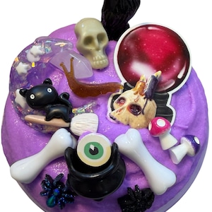 Può includere: Un oggetto decorativo rotondo viola con abbellimenti a tema Halloween. Presenta un corvo nero, un teschio, un gatto, un osso, un calderone e funghi. L'oggetto ha una finitura lucida ed è decorato con vari elementi spettrali.