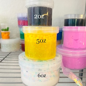 May include: Three clear plastic containers stacked on top of each other, filled with different colored slime. The top container is filled with black slime and labeled "2 oz", the middle container is filled with yellow slime and labeled "5 oz", and the bottom container is filled with white slime and labeled "6 oz".