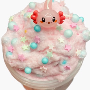 May include: A container of pink slime decorated with pastel-colored star-shaped sprinkles and small blue and white spheres. A pink axolotl figurine with red gills sits atop the slime. The slime has a fluffy, cloud-like texture.