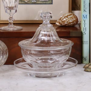 Peut inclure: Un bonbonnière en verre transparent avec un couvercle, posée sur une soucoupe assortie. Le plat présente un motif décoratif nervuré et un bouton sur le dessus du couvercle. Le plat est exposé sur une surface blanche, avec d'autres objets anciens en arrière-plan.