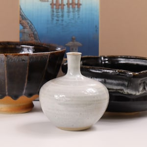 Könnte beinhalten: Eine Sammlung handgefertigter Keramik. Im Vordergrund steht eine kleine, cremefarbene Vase mit rundem Körper und schmalem Hals. Zwei dunkelbraune Schalen mit strukturierter Oberfläche und ein japanischer Druck im Hintergrund ergänzen das Arrangement.