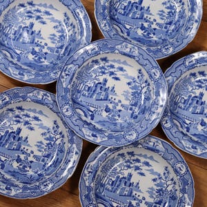 May include: Six blue and white porcelain plates with a landscape design. Each plate features a detailed scene of a castle, trees, and a bridge. The plates have a scalloped edge and are arranged on a wooden surface.