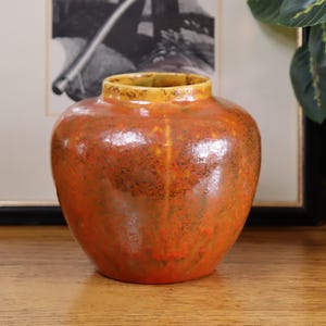 Puede incluir: Un jarrón redondo de cerámica con un esmalte moteado naranja y marrón. El borde es de color amarillo contrastante. El jarrón está sobre una superficie de madera, con una imagen enmarcada y vegetación en el fondo.